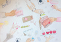 A square white art print or poster featuring a watercolor collage of illustrated hands holding cocktails, cherries, olives, playing cards, and a pink drink, laid flat on a light wood floor. The word 'Santor' appears in script at the top right.