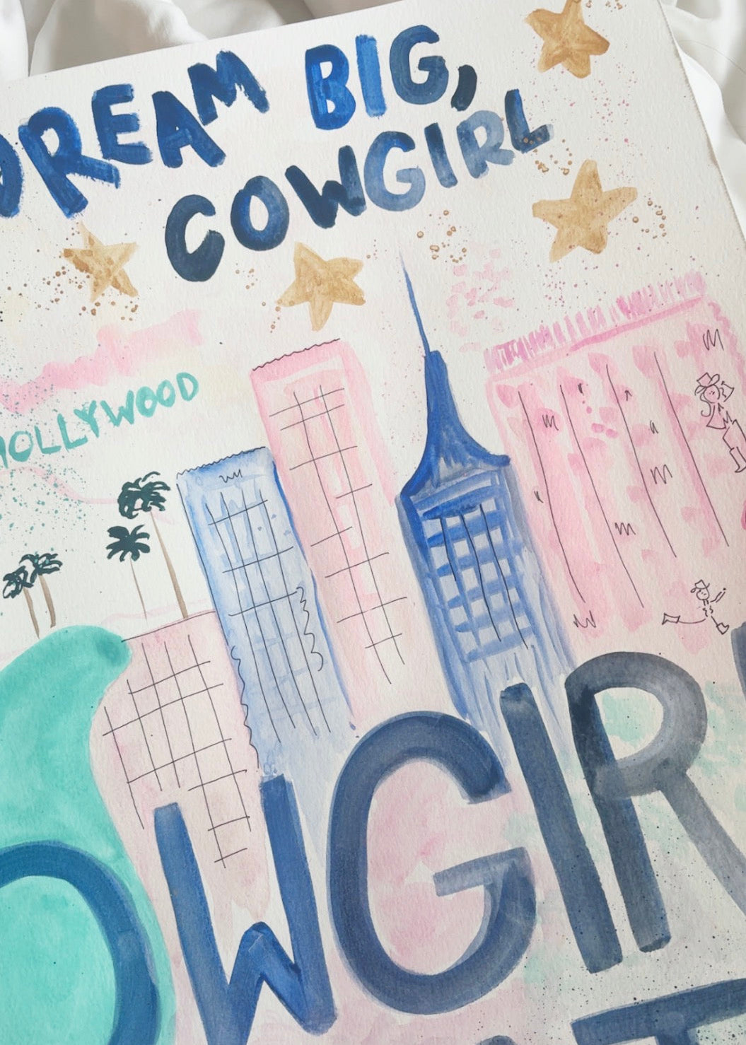 A close-up of a hand-painted poster or art print featuring the text 'DREAM BIG, COWGIRL' and 'COWGIRL CUTIE' in blue and pink lettering, with watercolor-style buildings, stars, and a teal shape on a white background.