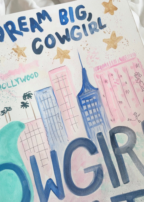 A close-up of a hand-painted poster or art print featuring the text 'DREAM BIG, COWGIRL' and 'COWGIRL CUTIE' in blue and pink lettering, with watercolor-style buildings, stars, and a teal shape on a white background.